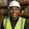 Professional portrait of a smiling warehouse manager in safety gear. Headshot of an African American man in a warehouse wearing a white hard hat, yellow safety vest, and dark shirt, smiling in front of stacked storage racks.