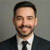 Business portrait of a cheerful male professional. Bearded man with short dark hair, smiling warmly in a suit and tie with a neutral gray background.