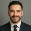 Business portrait of a cheerful male professional. Bearded man with short dark hair, smiling warmly in a suit and tie with a neutral gray background.